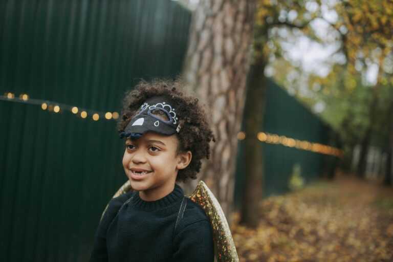 Young girl in Halloween costume and mask smiling outdoors among trees in autumn.