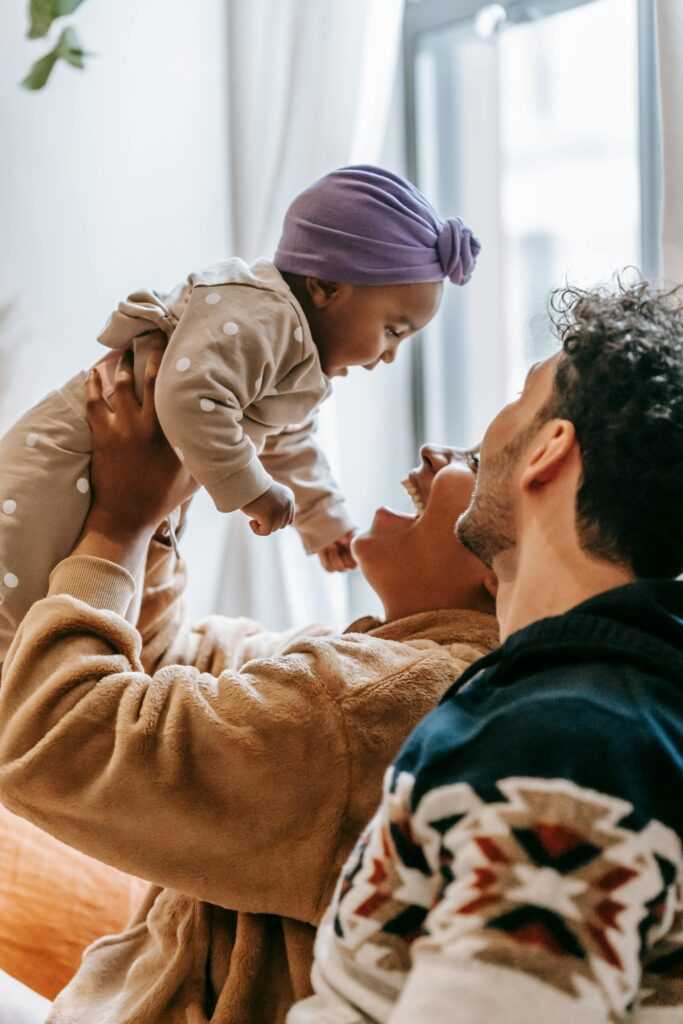 Side view of cheerful ethnic mother lifting cute baby girl while sitting near positive father at home