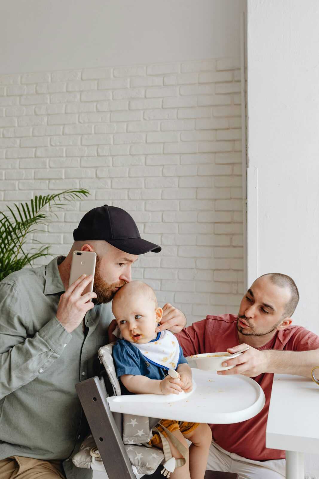 Two fathers with a baby in a high chair, sharing a caring moment in a bright kitchen.